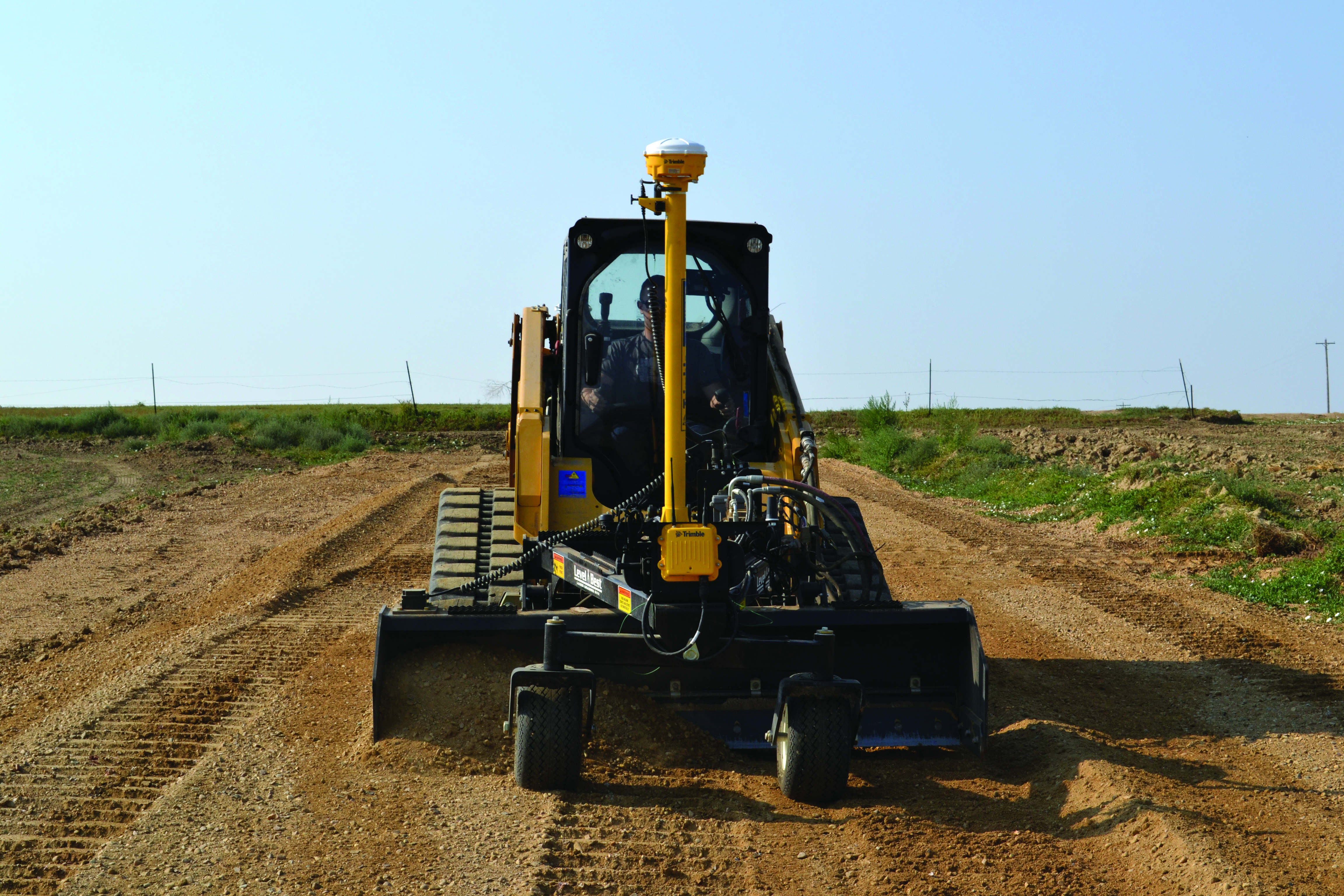 El sistema 3D Trimble Earthworks equipa un cargador compacto Bobcat con accesorio de nivelación en un entorno de construcción. La avanzada tecnología GPS en los postes amarillos es visible, lo que indica una mayor capacidad de precisión para una mayor productividad y eficiencia en la preparación del terreno