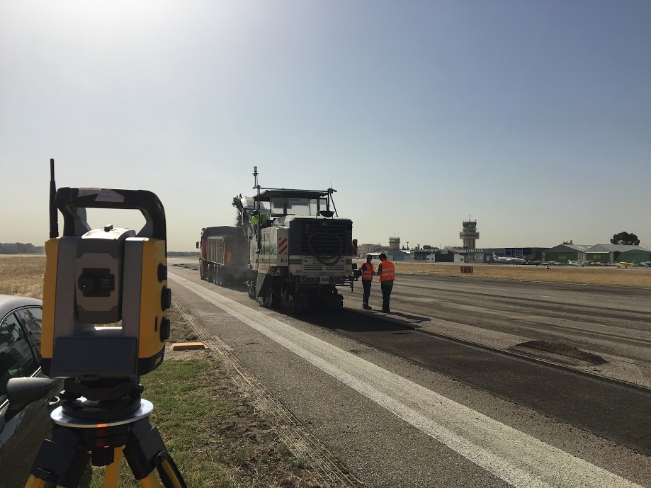 Estação total topográfica em primeiro plano utilizada em sistemas de orientação de fresadoras em obras rodoviárias. Operadores que trabalham com máquinas de pavimentação numa estrada em construção