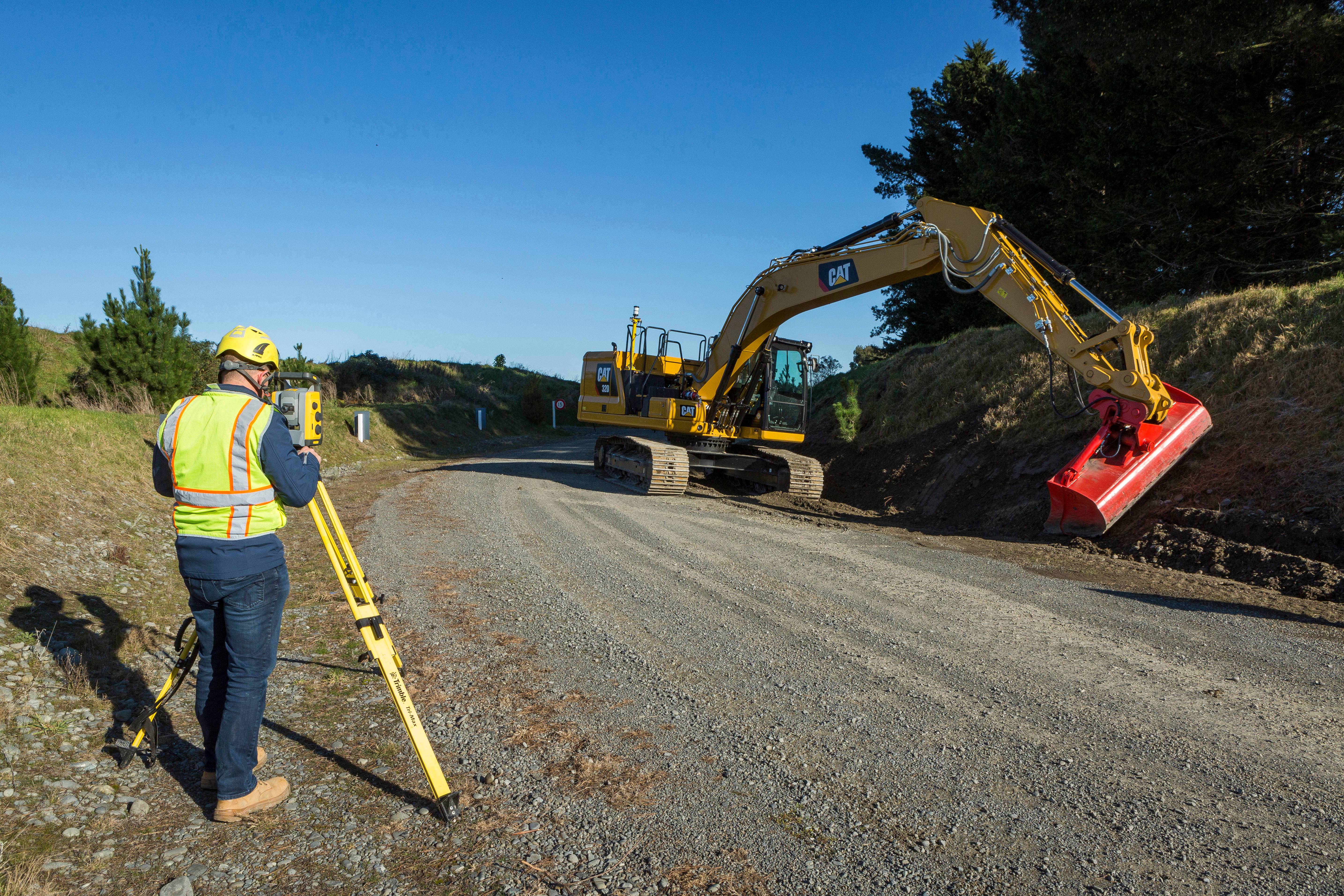 Operario trabaja en la configuración de un sistema de nivelación utilizando una estación total, mientras una excavadora amarilla realiza trabajos en una carretera de grava.